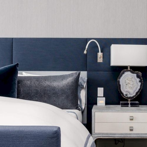 A modern bedroom with a blue headboard, a bed with pillows, and a nightstand holding a lamp, book, and phone.